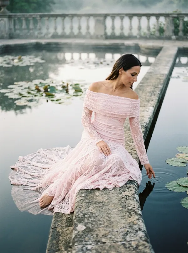 the same blush lace gown seated on the stone edge of a lily pond at first light